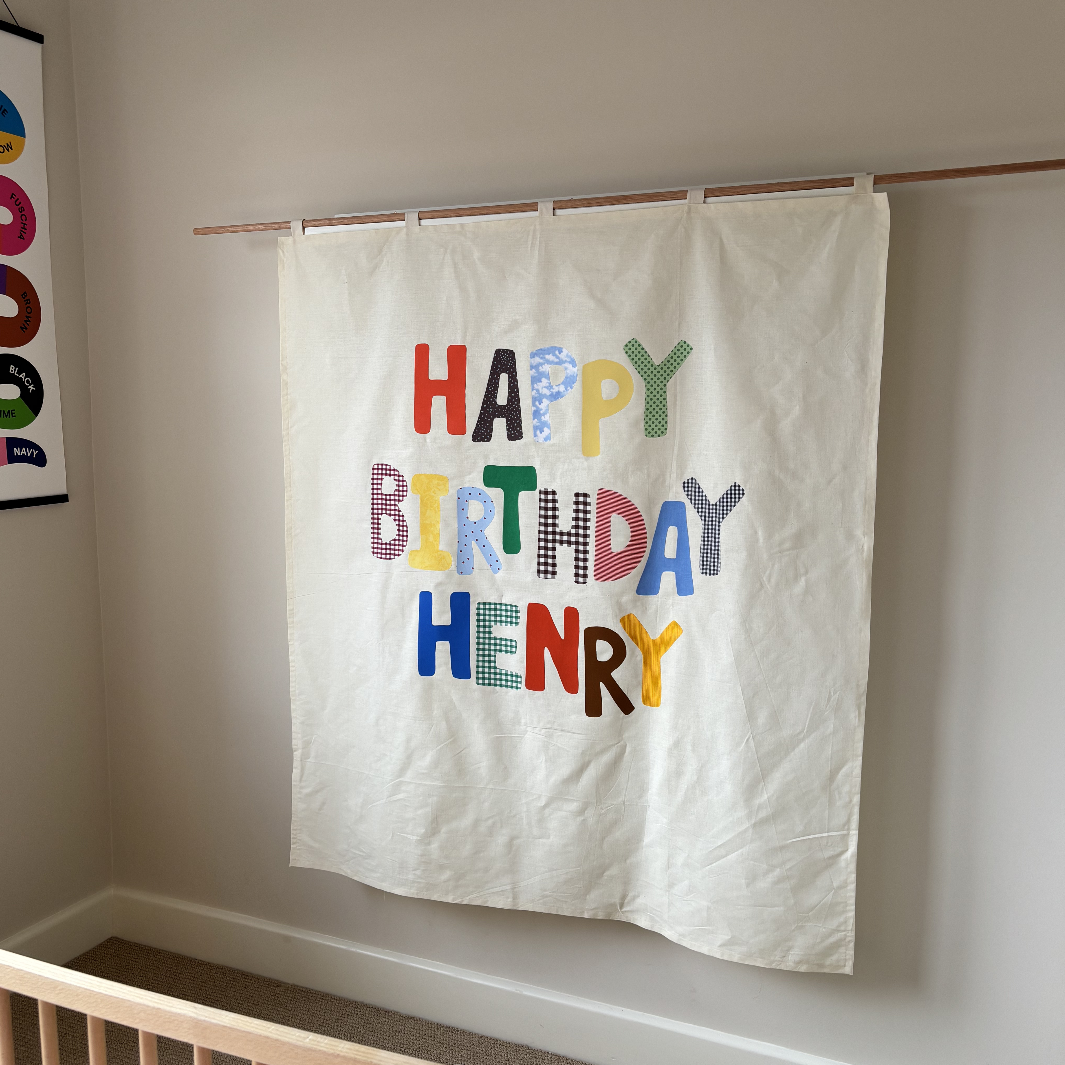 Handmade birthday banner with colourful fabric letters appliquéd onto calico spelling out HAPPY BIRTHDAY HENRY hanging from a wooden rod on a cream background. Made by Bugalugs in Melbourne.