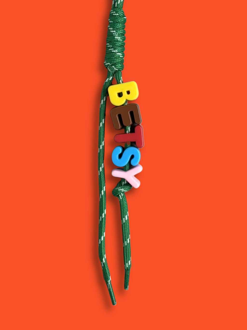 Personalised bag charm with letter beads spelling BETSY in yellow, brown, red, blue and pink, threaded on a green and white fleck knotted cord, with a purple carabiner clip on a red background. Made by Bugalugs in Melbourne.