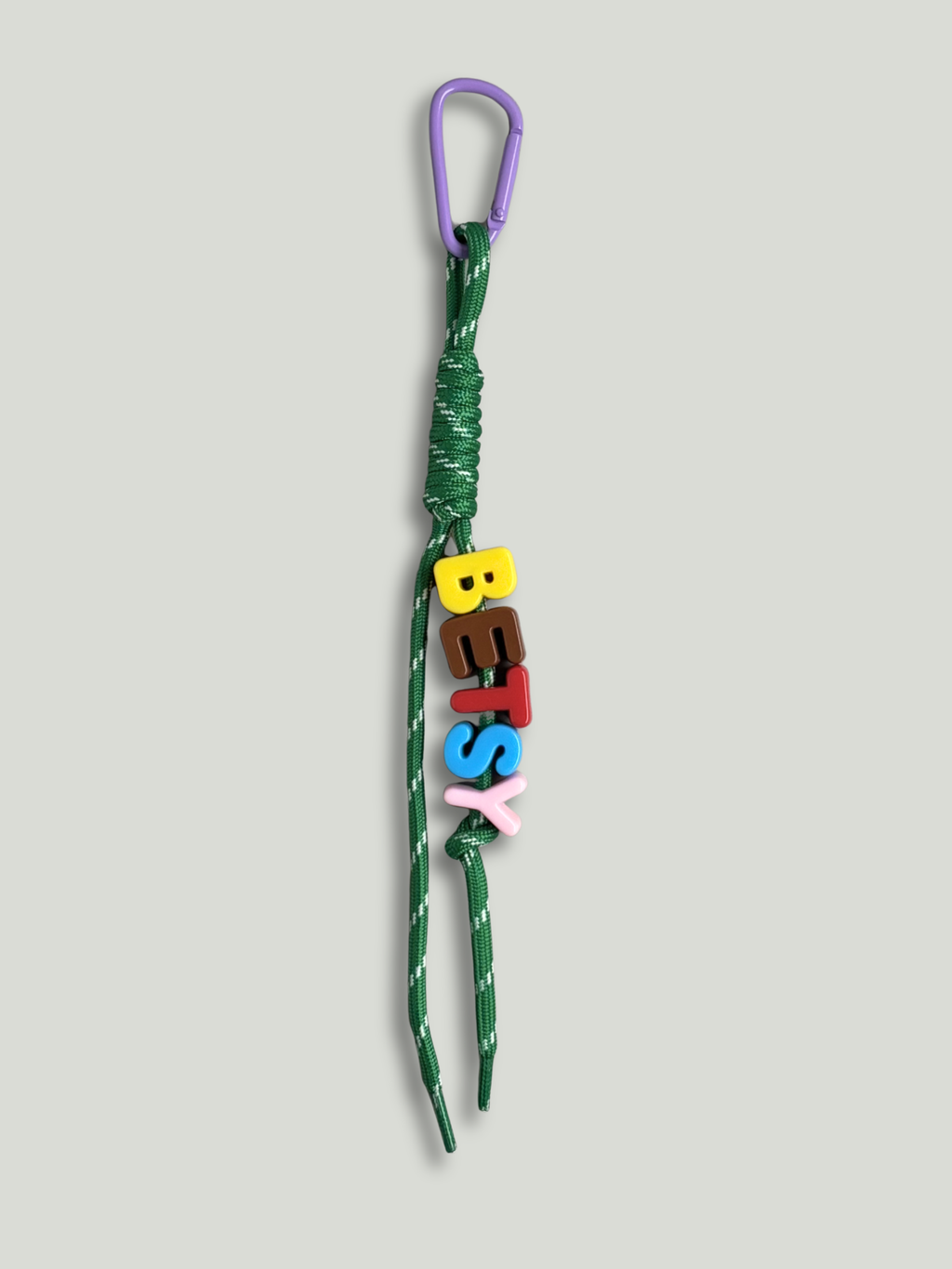 Personalised bag charm with letter beads spelling BETSY in yellow, brown, red, blue and pink, threaded on a green and white fleck knotted cord, with a purple carabiner clip on a yellow background. Made by Bugalugs in Melbourne.