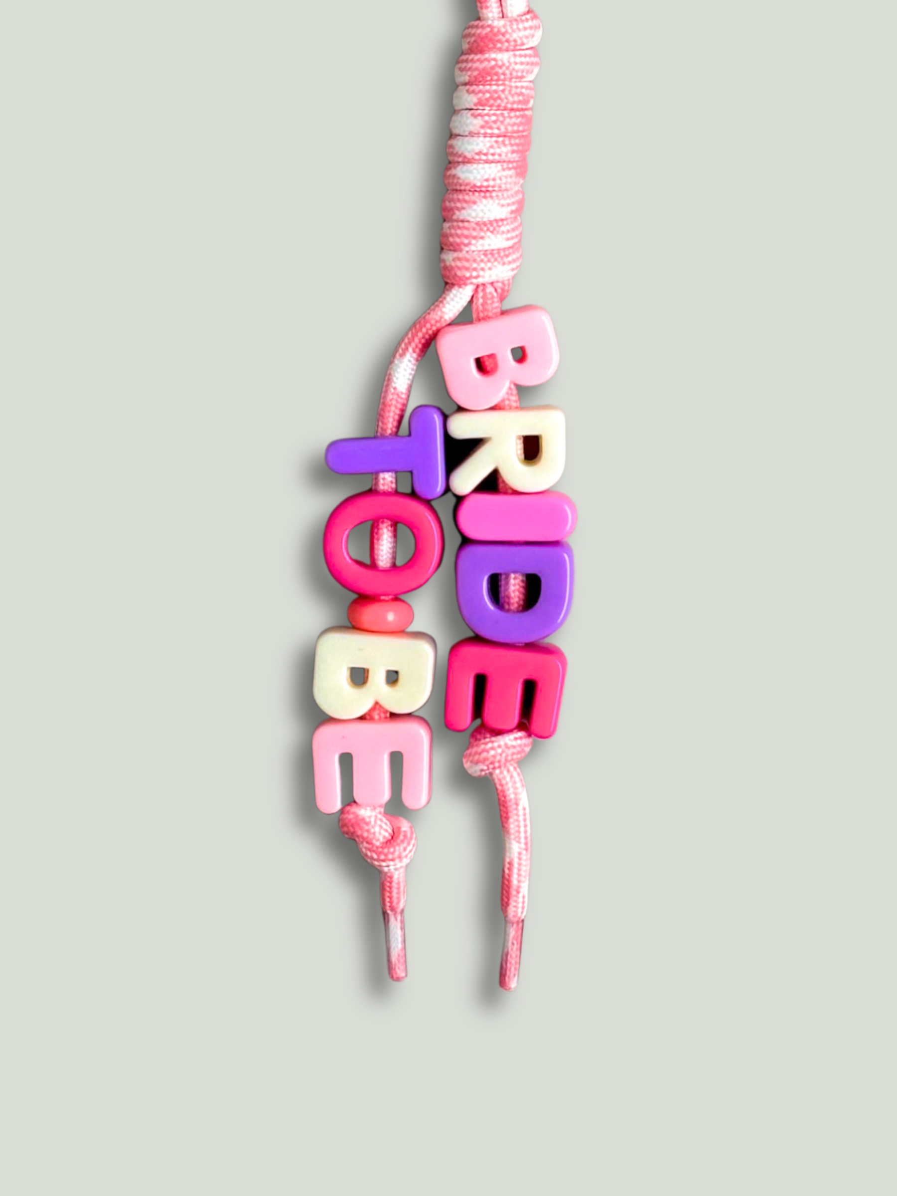 Custom statement bag charm with letter beads spelling BRIDE TO BE in pink, white and purple, threaded across two strands of a pink and white houndstooth knotted cord with a purple carabiner clip, on a gray background. Made by Bugalugs in Melbourne.