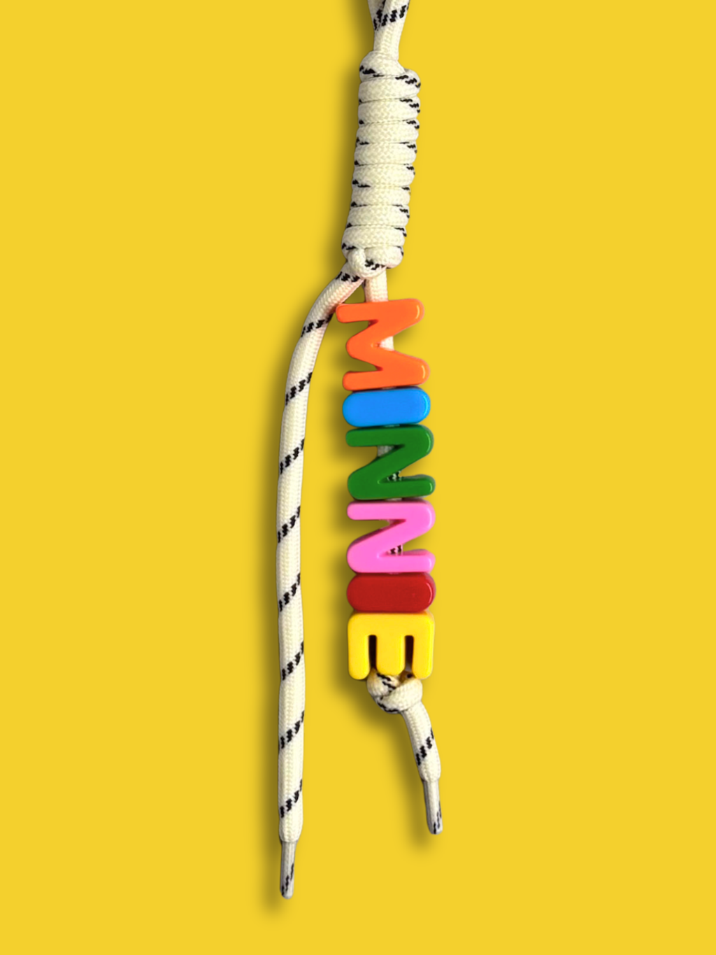 Personalised bag charm with letter beads spelling MINNIE in orange, blue, green, pink, red and yellow, threaded on a cream and black fleck knotted cord, with a red carabiner clip on a yellow background. Made by Bugalugs in Melbourne.
