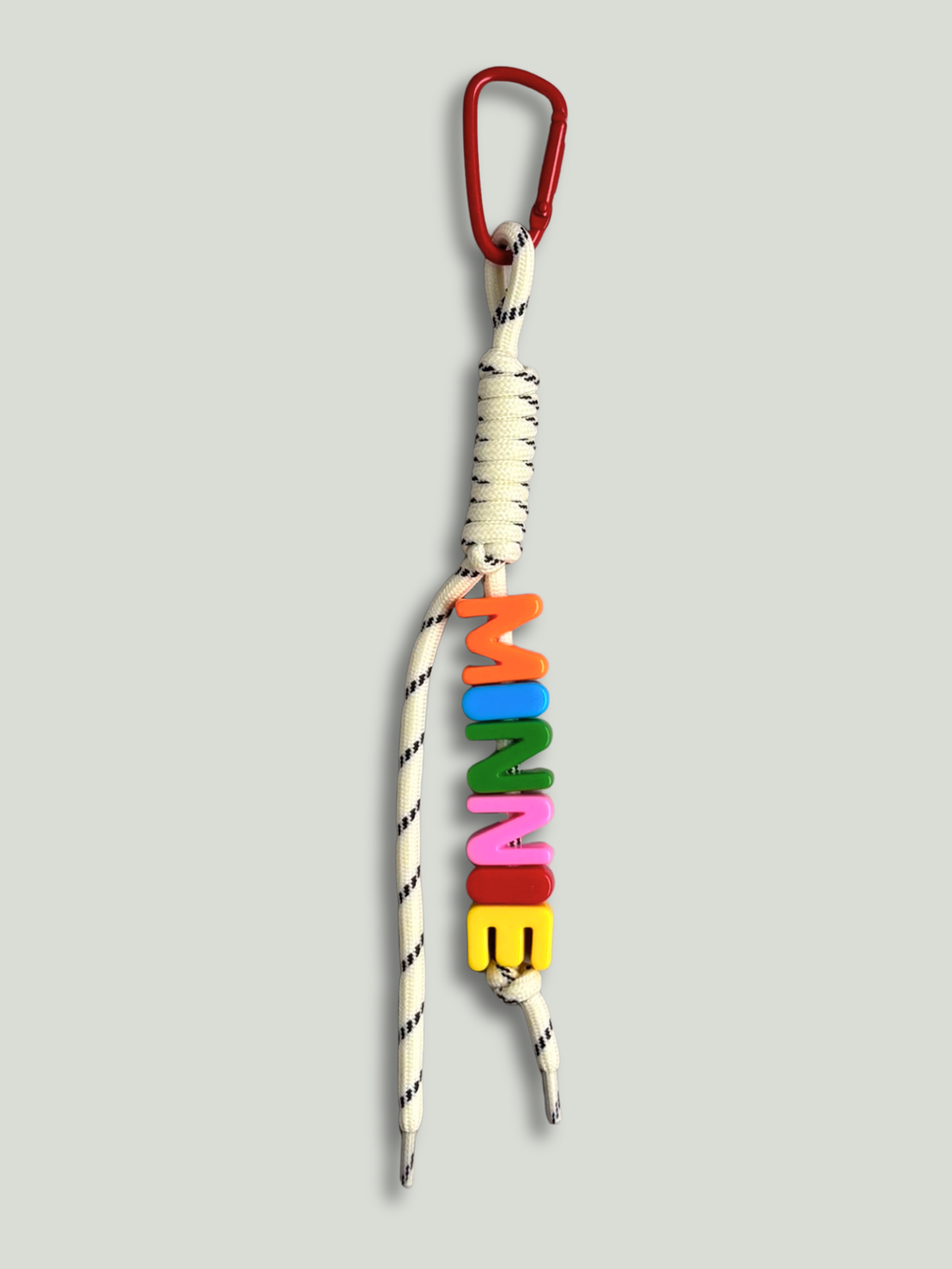 Personalised bag charm with letter beads spelling MINNIE in orange, blue, green, pink, red and yellow, threaded on a cream and black fleck knotted cord, with a red carabiner clip on a gray background. Made by Bugalugs in Melbourne.