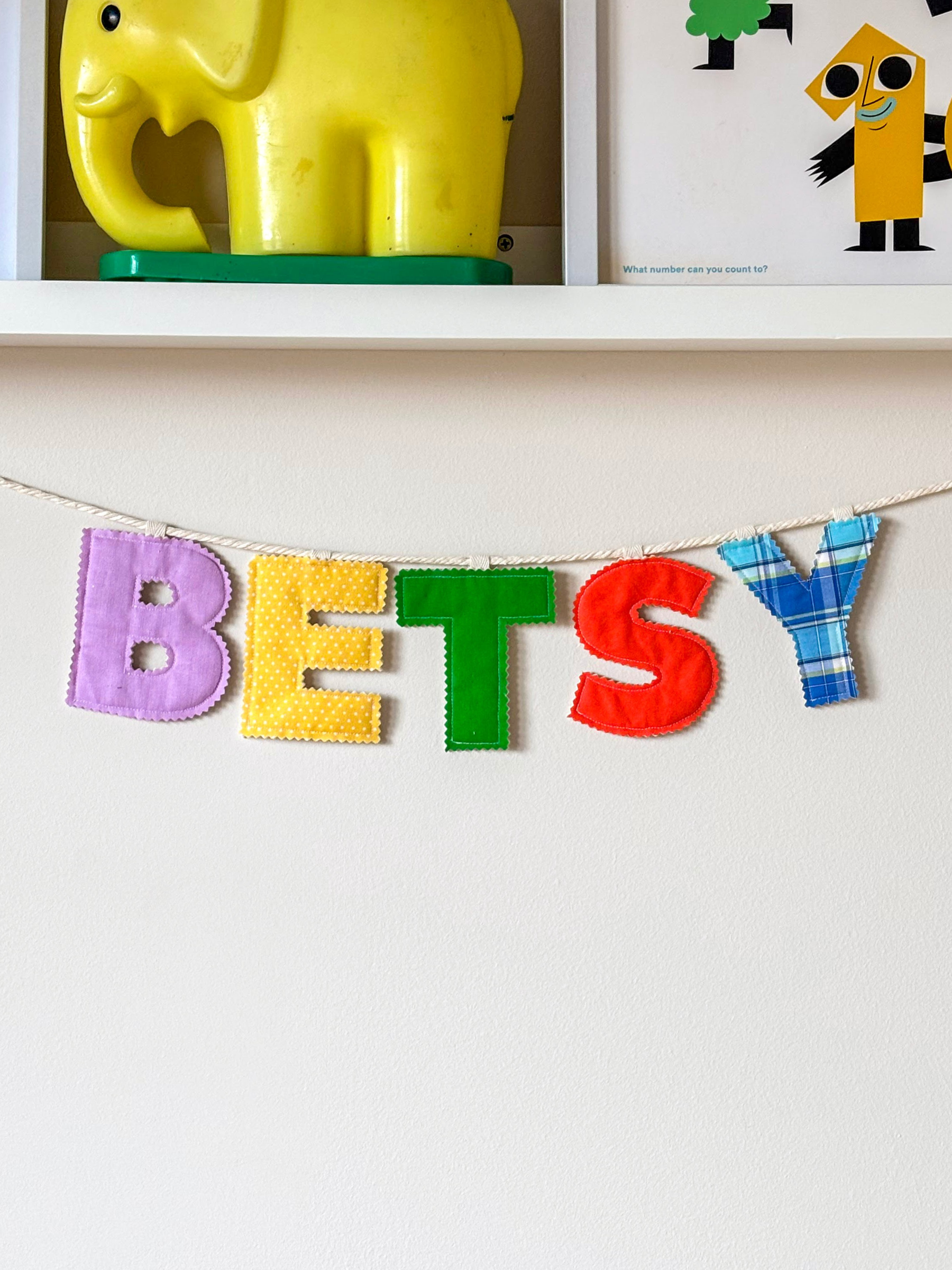 Personalised name banner spelling 'BETSY' in bright fabrics on a white wall. Made by Bugalugs in Melbourne. 