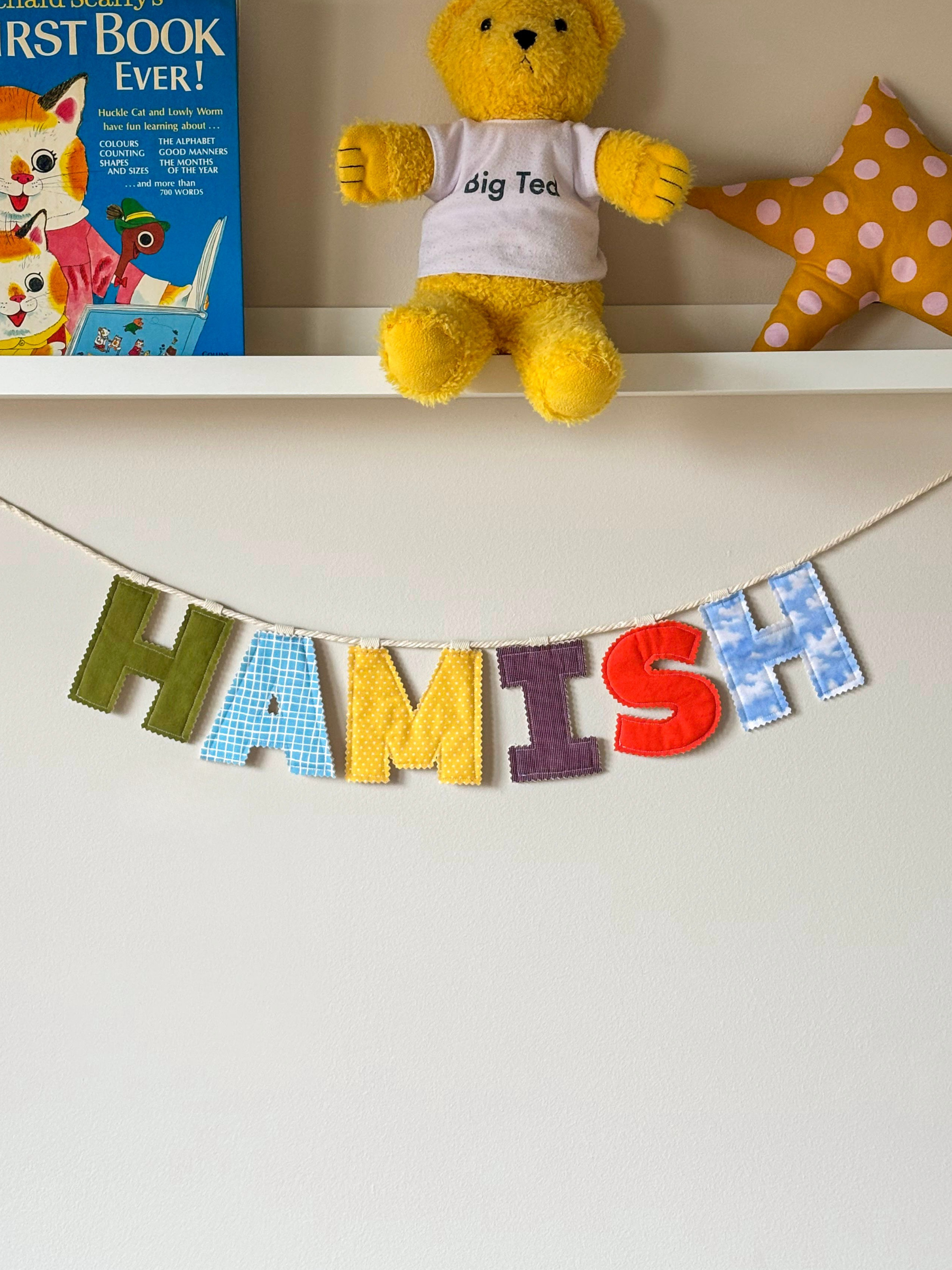 Personalised name banner spelling 'HAMISH' in bright fabrics on a white wall. Made by Bugalugs in Melbourne. 